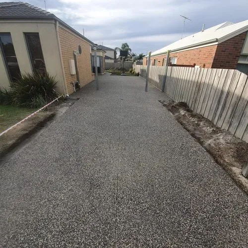 Concrete driveway with a gravel texture, flanked by houses and a wooden fence.